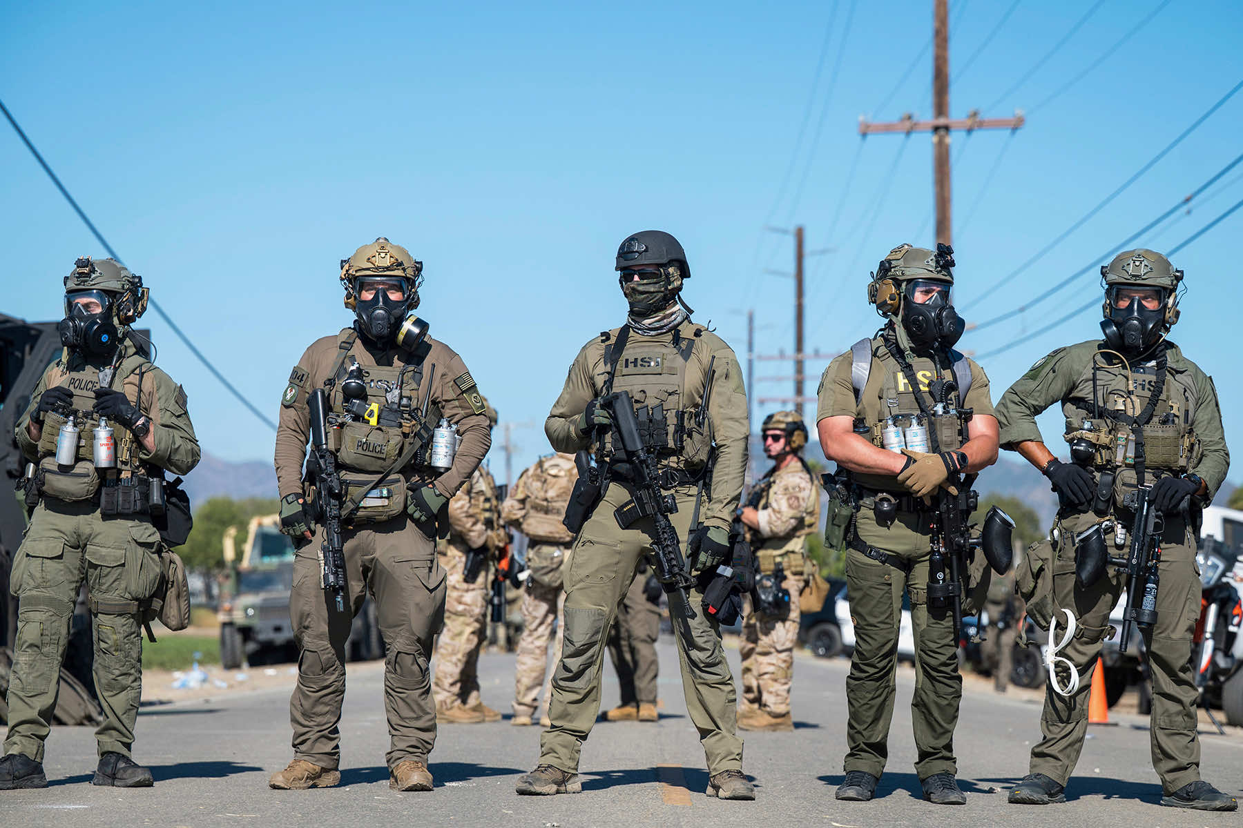 Five ICE HSI agents in olive tactical gear with gas masks and rifles standing in formation on a road with desert mountains behind them