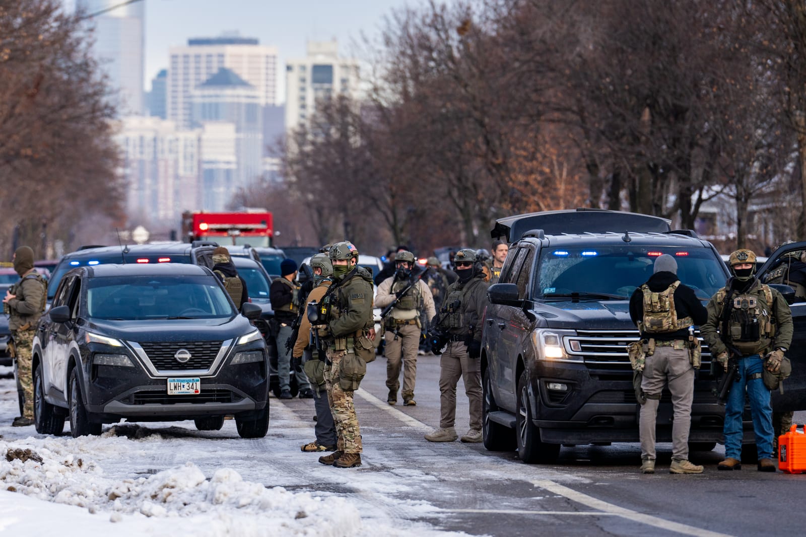 Multiple ICE agents in tactical gear conducting a traffic stop on a snowy urban street with a city skyline in the background