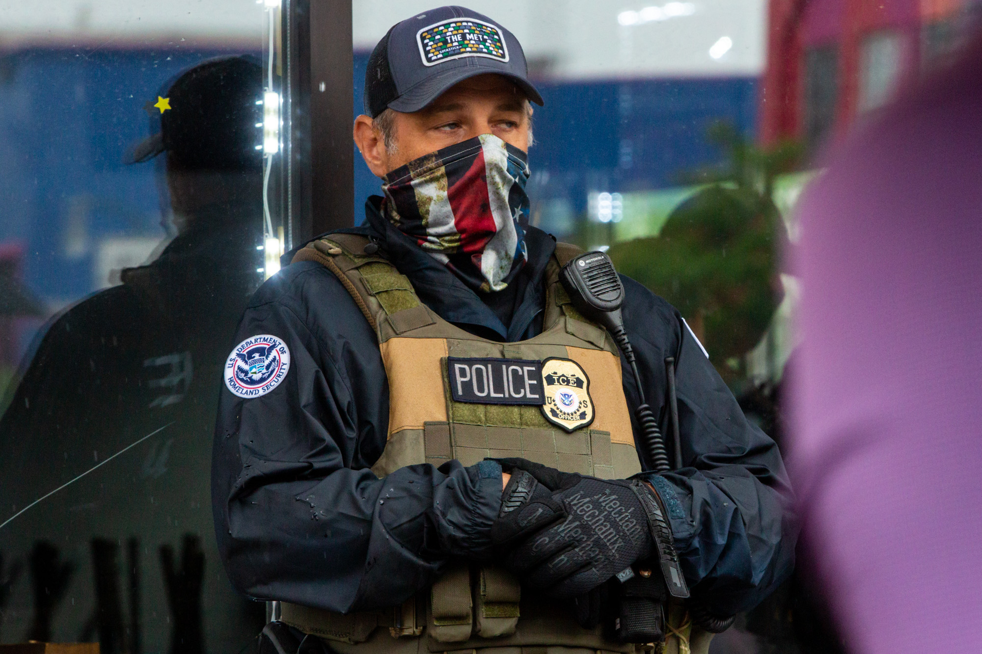 Close-up of an ICE agent wearing a Department of Homeland Security shoulder patch, POLICE vest with ICE badge, and an American flag face covering
